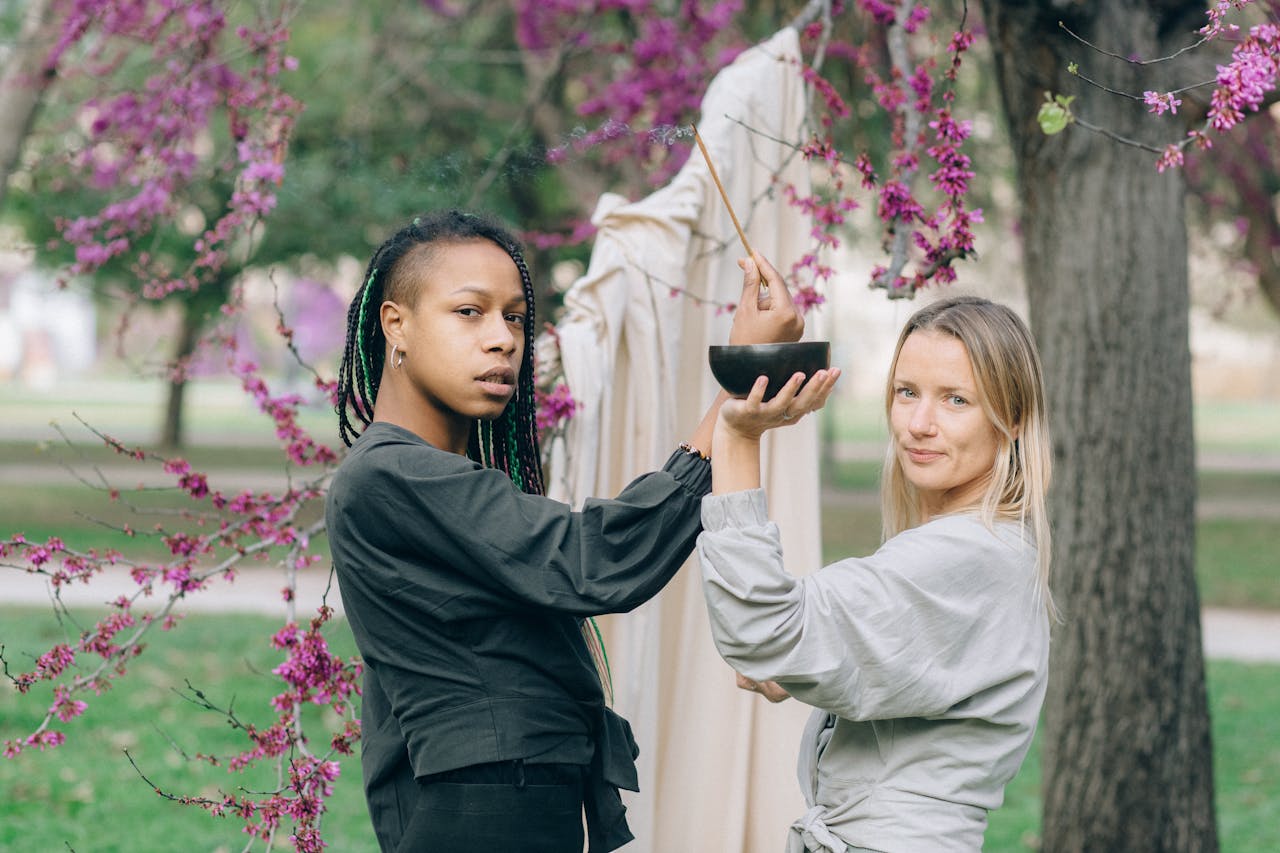 Two women holding an incense bowl surrounded by spring blossoms outdoors.